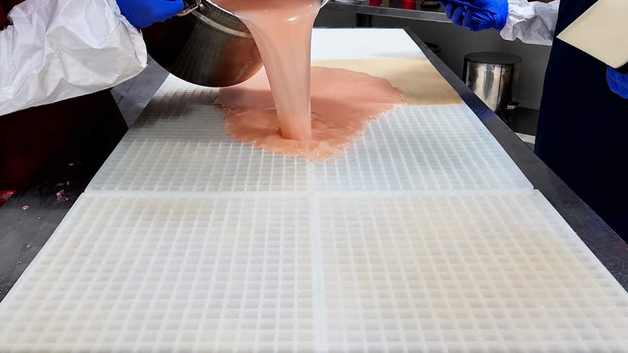 Echipa Gummy Molds toarnă amestecul pentru jeleuri peste o formă comercială mare din silicon înainte de răzuire